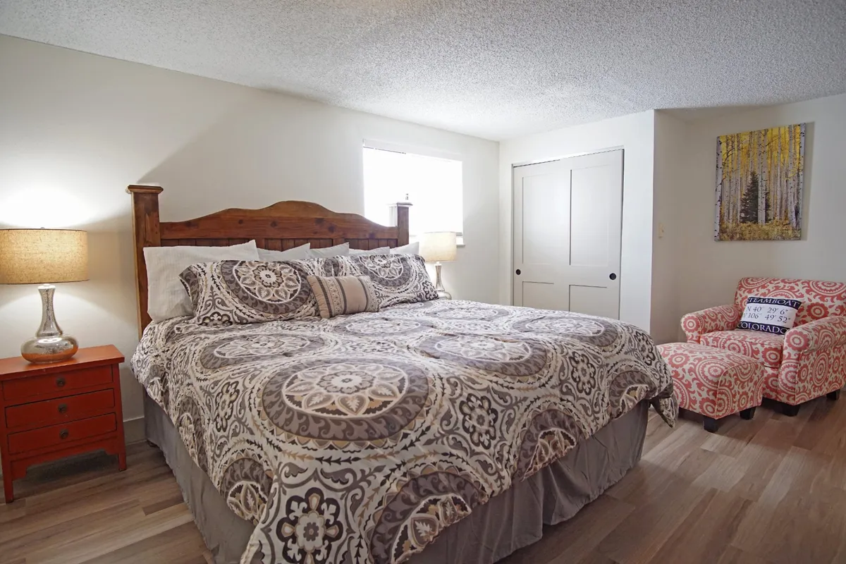 A spacious bedroom featuring a king-size bed, a comfortable armchair with ottoman set beside a lamp for reading, large windows letting in natural sunlight, and tasteful artwork on the walls. The setting feels private and tranquil, offering a cozy corner perfect for relaxation.