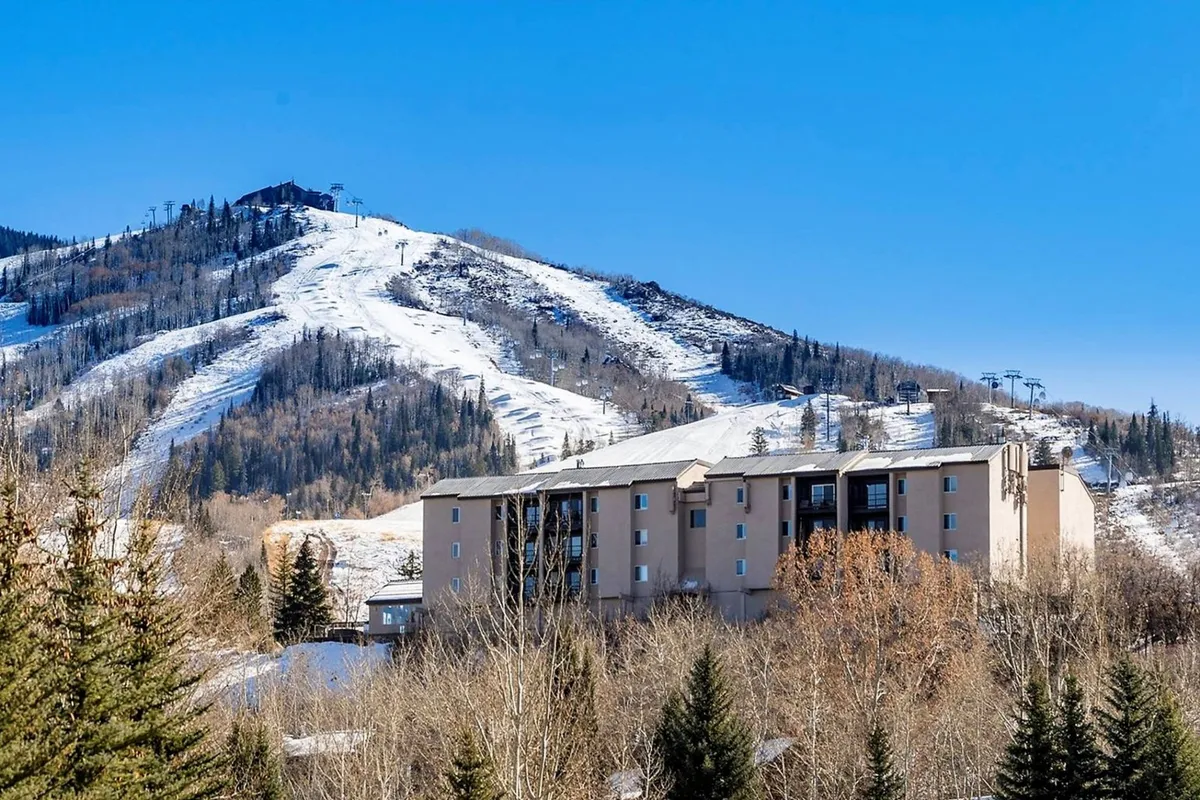 An exterior shot featuring the property’s building set against a striking backdrop of a snow-capped mountain and lush trees. While some snow remains, the prominence of trees and the hotel building conveys a sense of arrival and the property's relationship to the outdoors.