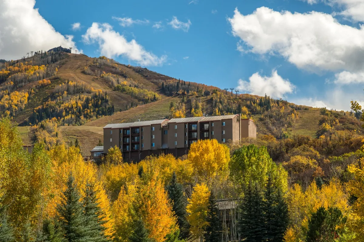 The exterior of the building is nestled among trees with vibrant autumn foliage in a mountainous setting, reflecting both secure surroundings and the condo’s elevated position for seasonal enjoyment. The inviting approach to the property connects to both practical access and natural beauty.