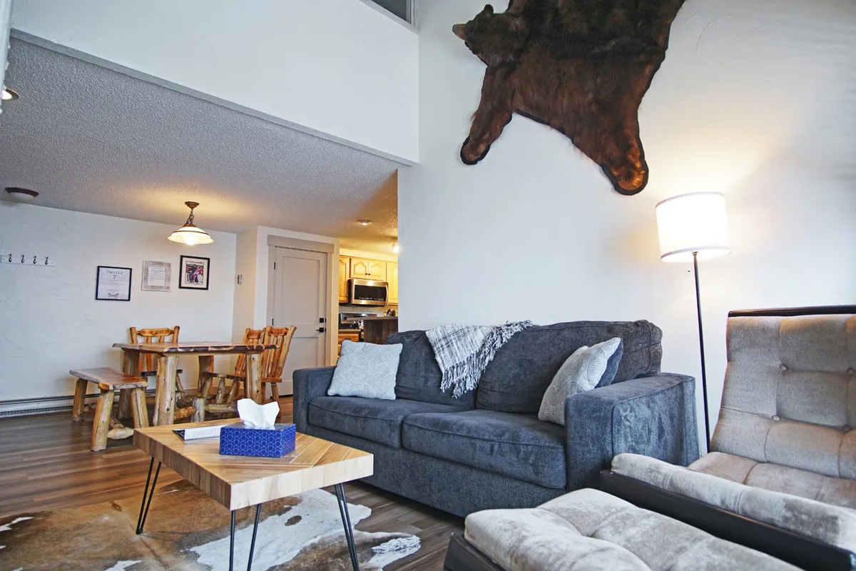 A warm, inviting living and dining area with a plush sofa, rustic wooden dining table, bear skin rug, and soft lighting. The open-concept layout is designed for social gatherings, with comfortable seating for family dinners or cozy movie nights, set against the glow of ambient lighting.
