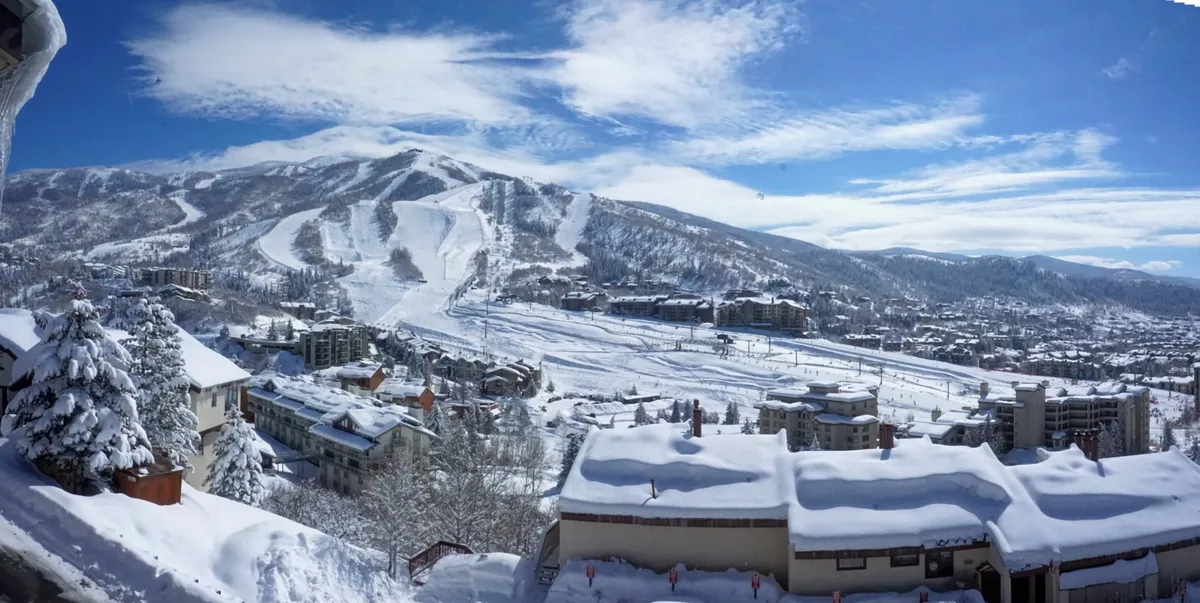 A panoramic view looking out over snow-blanketed mountains, ski runs, scattered residential buildings, and pine trees under a crisp sky. The image radiates both adventure and peace, hinting at the experiences awaiting guests both on and off the slopes.