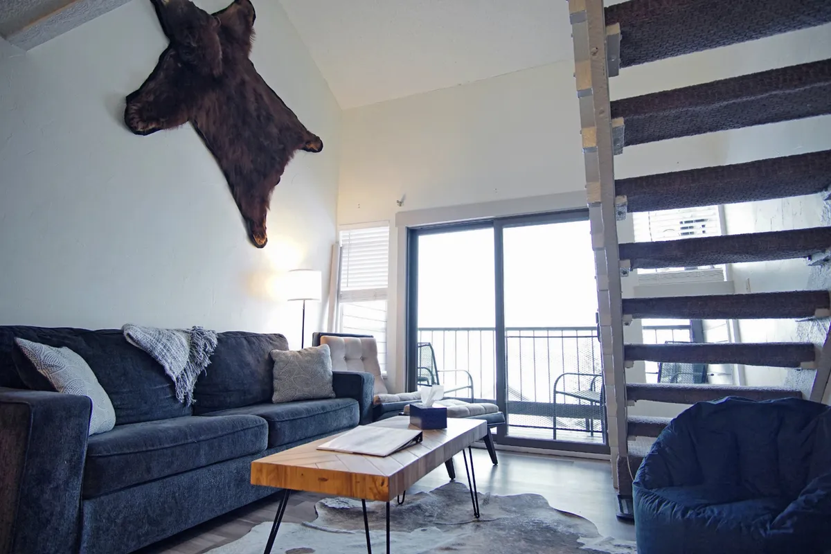 A cozy living area featuring a sofa, cowhide rug, wooden coffee table, staircase, and large windows letting in natural light. The space is ideal for socializing, reading, or simply relaxing, with a rustic yet comfortable ambience.