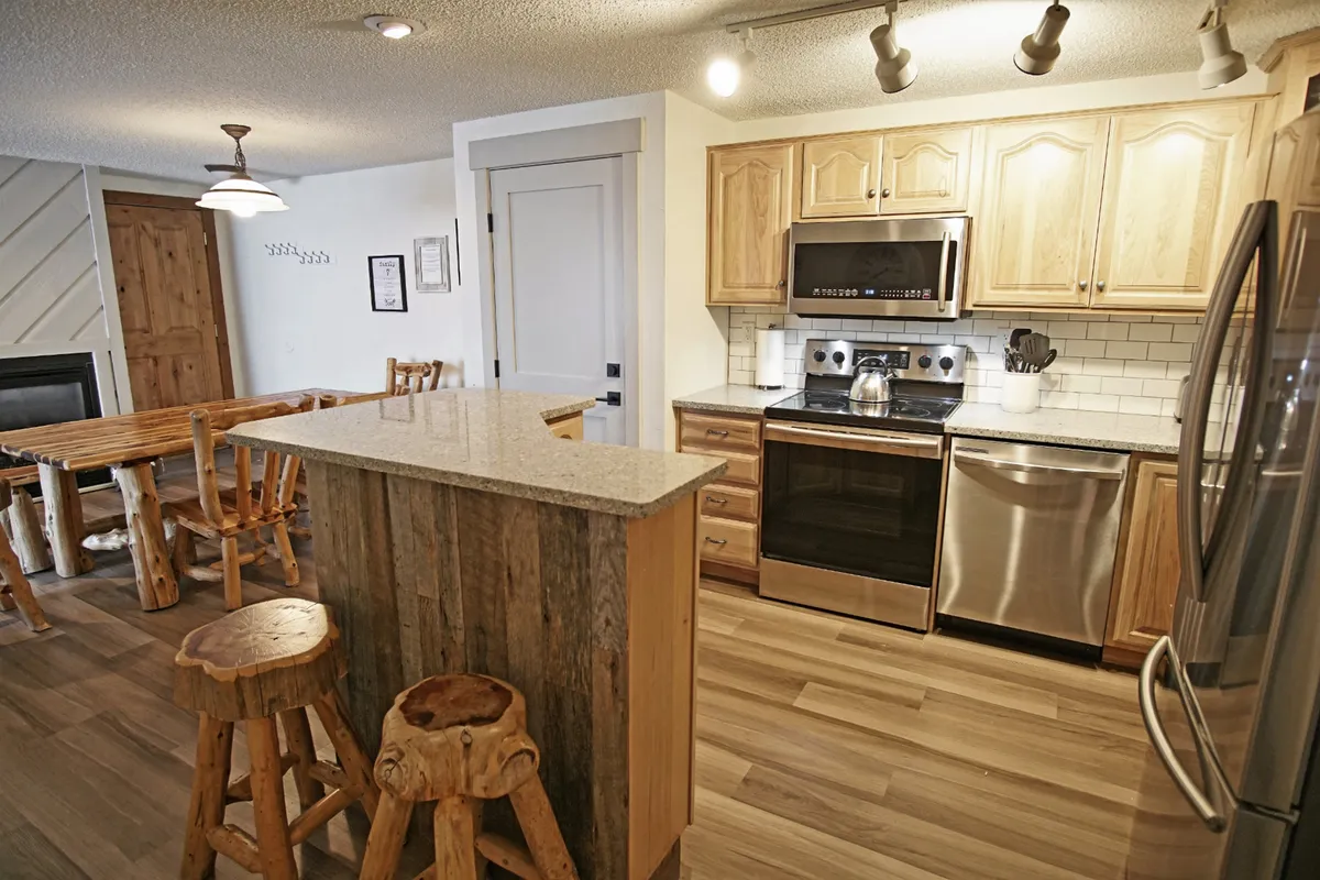 A view of the kitchen island with stainless steel appliances, a wooden dining table, and rustic stools, all enveloped in a warm, welcoming mood. The space is primed for cooking, entertaining, and casual family gatherings.