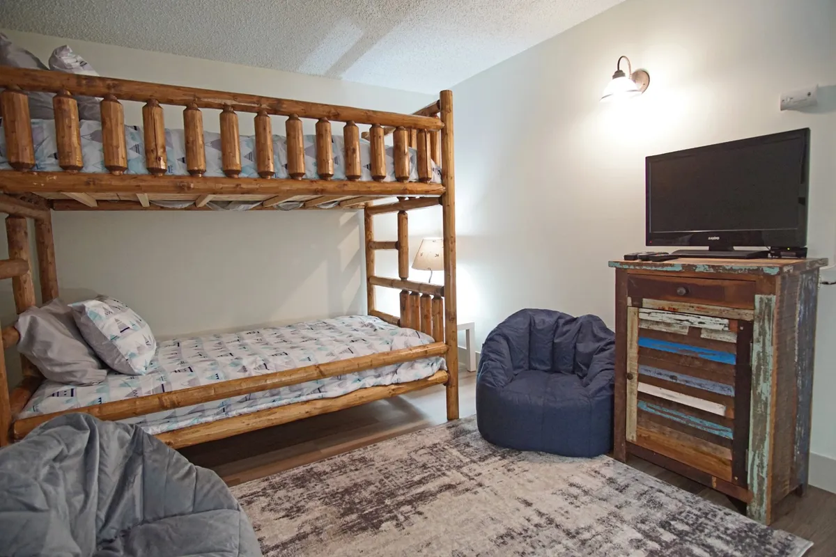 A bright loft area overlooking the living space below, featuring a bunk bed, a bean bag chair, a TV atop a wooden dresser, and inviting decor. The layout is semi-private, ideal for older children or friends seeking a unique sleep spot.