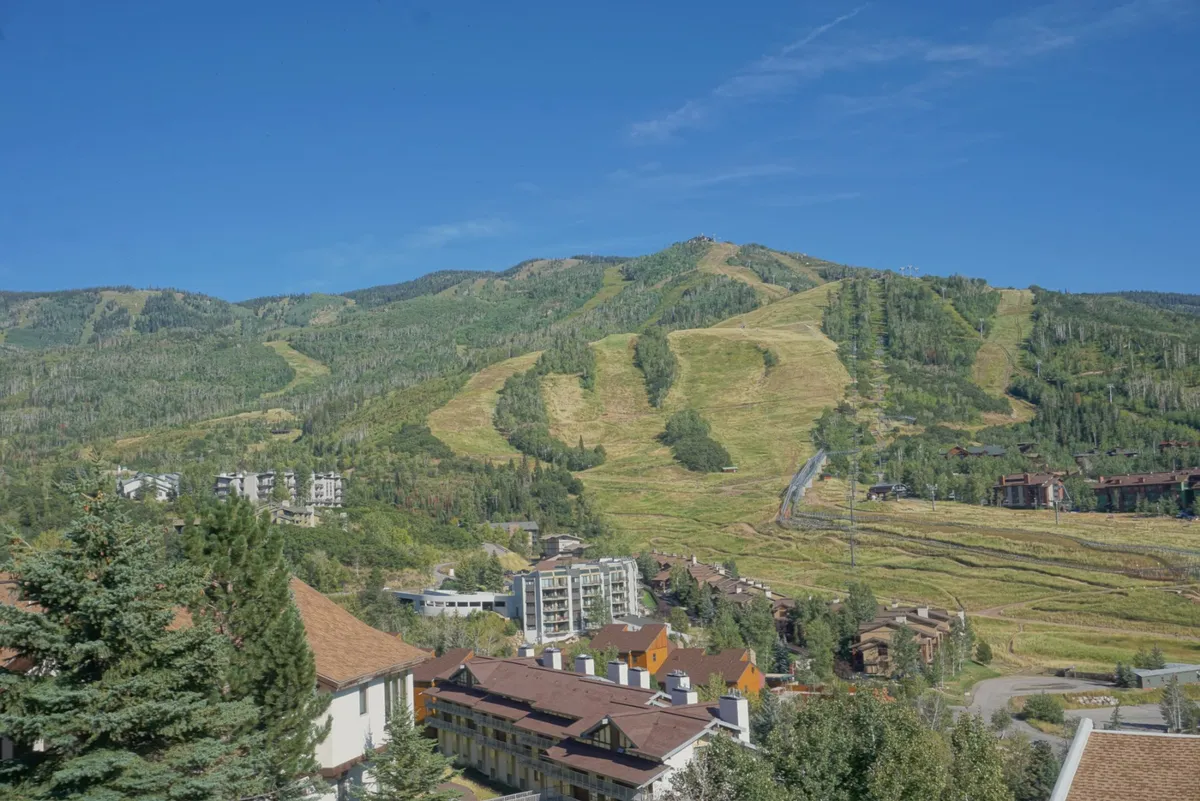 The image showcases the exterior view from the condo, with sweeping vistas of Steamboat Ski Resort’s pristine slopes, lush evergreen trees, and charming buildings nestled in the foreground, all set beneath a vibrant, clear blue sky. The scene is tranquil yet invigorating, highlighting the stunning mountain landscape and proximity to outdoor adventure.
