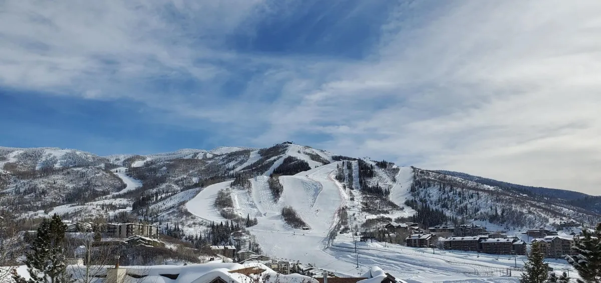 A sweeping winter exterior showcasing snow-covered slopes, a mountain range in the distance, active ski lifts, and residential buildings nestled among frosted trees. The scene conveys both the excitement of outdoor activities and the tranquil beauty of Steamboat's mountain setting.