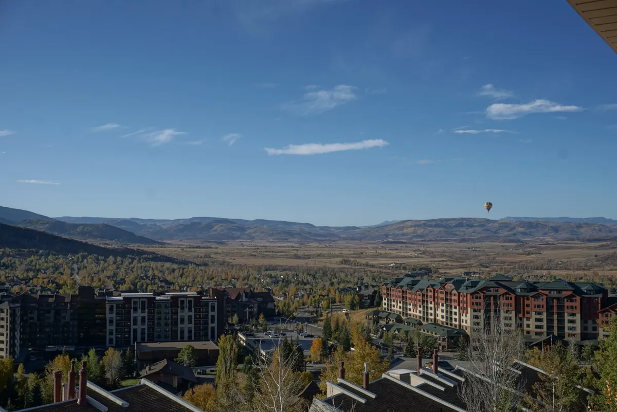 A panoramic view from the balcony reveals a stunning mountain range, colorful fall foliage, and a hot air balloon rising at dawn. The scene conveys a sense of peaceful adventure and highlights the connection to the outdoors.