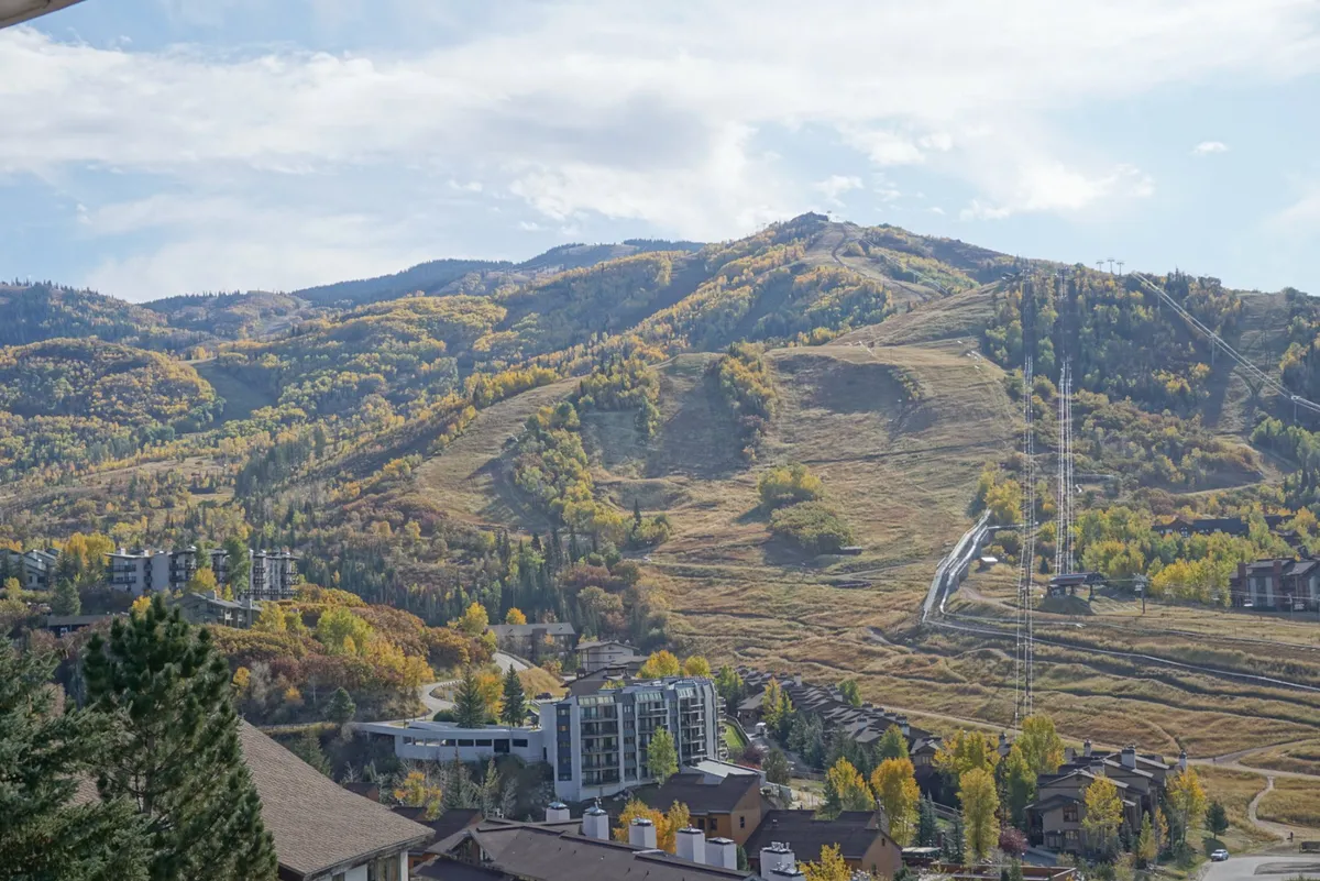 This view image captures a breathtaking panorama of the surrounding mountain range, lush green tree lines, and the valley below. Residential buildings nestle at the base, hinting at the property's proximity to outdoor adventure. The scene is tranquil but filled with the promise of exploration and activity.