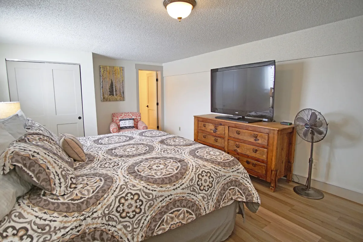 A restful queen bedroom with patterned bedding, a wooden dresser topped with a television, a ceiling fan, and an accent chair in the corner. The room exudes an inviting atmosphere, with soft lighting that highlights its intimate and comfortable setting.
