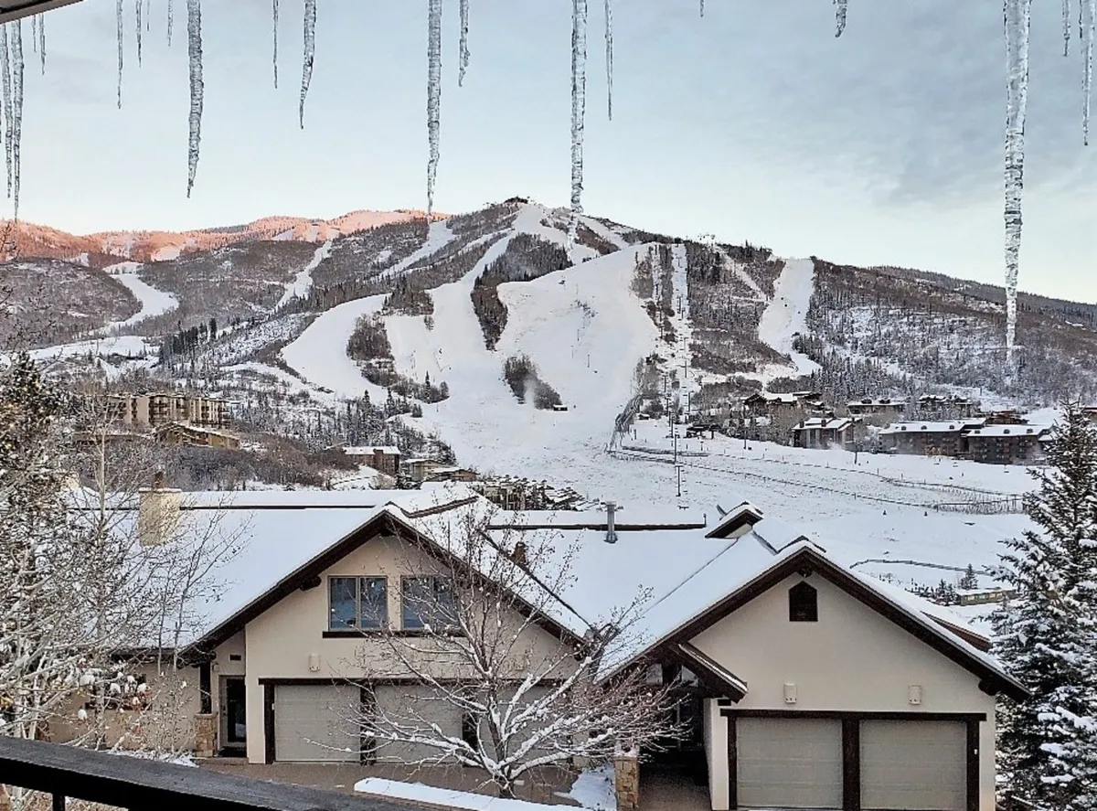 This exterior shot captures snow-covered mountains and ski slopes, with icicles hanging from the roof and nearby homes visible. The mood is both tranquil and adventurous, offering a sense of the area's winter wonderland and proximity to outdoor activities.