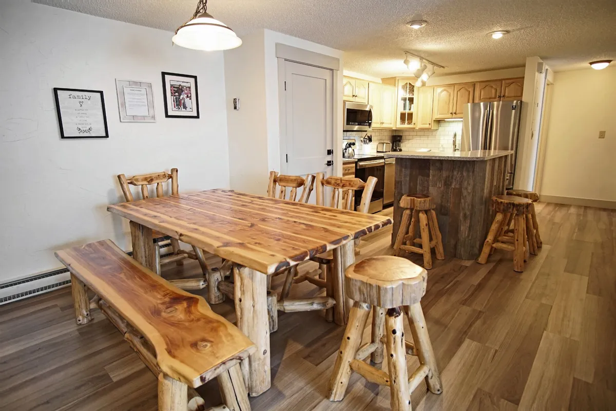 A close look at the rustic wooden dining table paired with natural finish stools and modern kitchen appliances nearby. Family-themed wall art enhances the cozy and inviting atmosphere, emphasizing the space's suitability for meals, games, and celebrations.